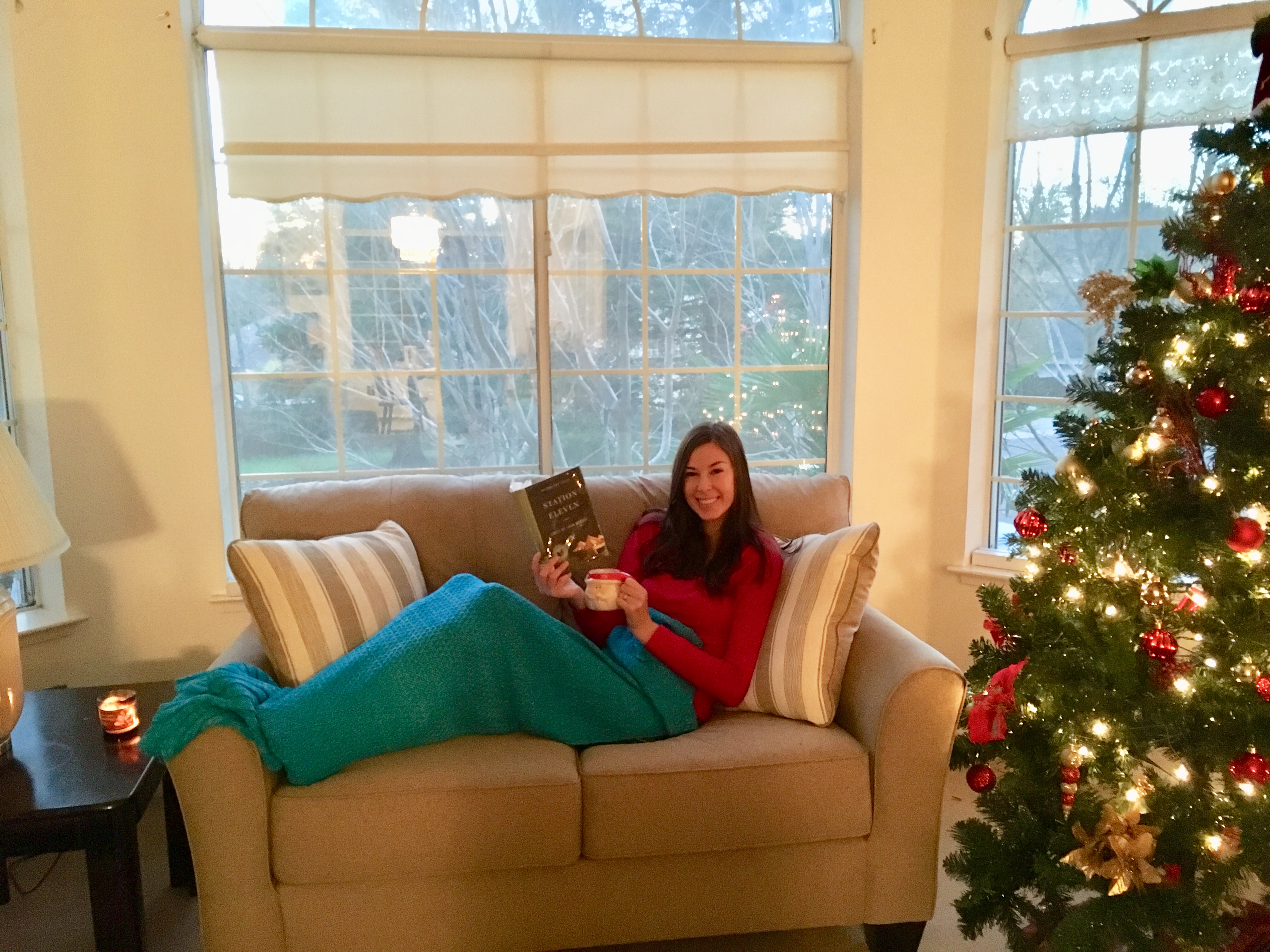 Jenna sitting on couch with mug and book in a teal blanket