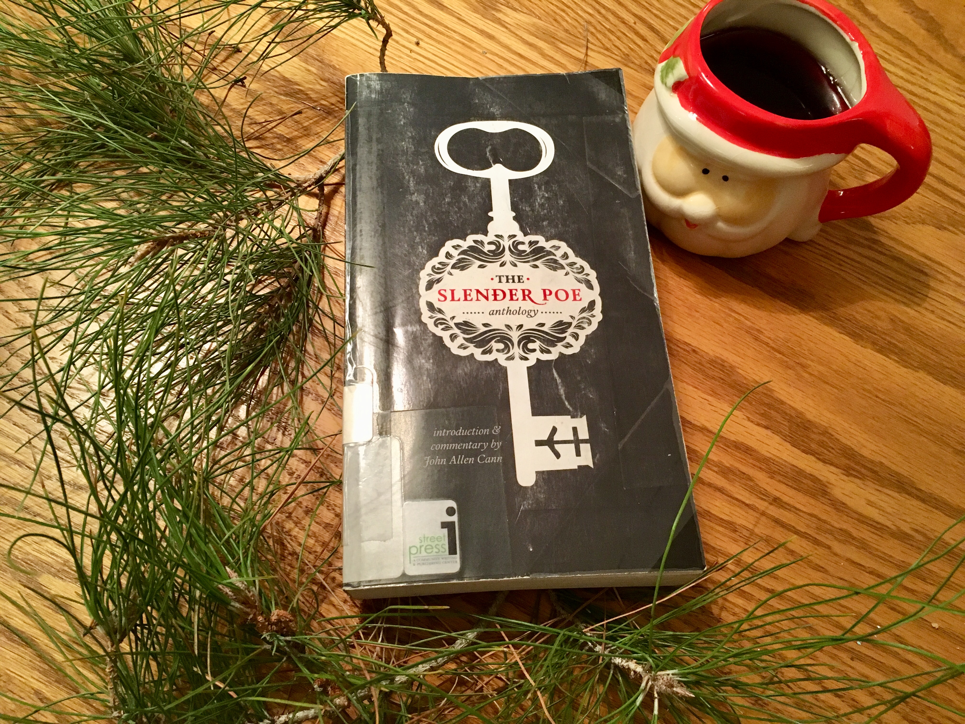 The Slender Poe Anthology paperback on wood table next to pine needles and coffee in Santa mug