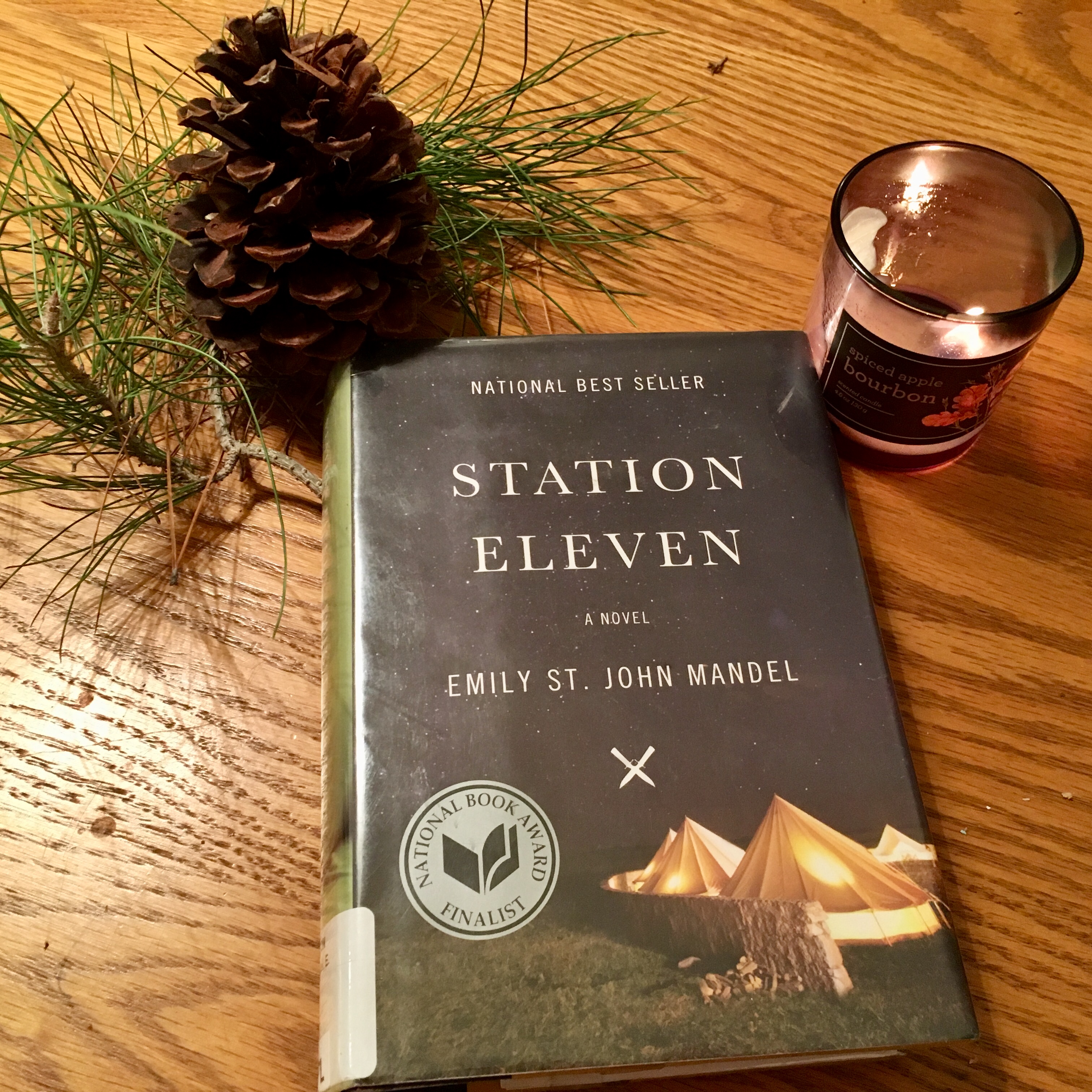 Station Eleven hardback sitting on wood table next to pine needles, a pine cone, and a candle