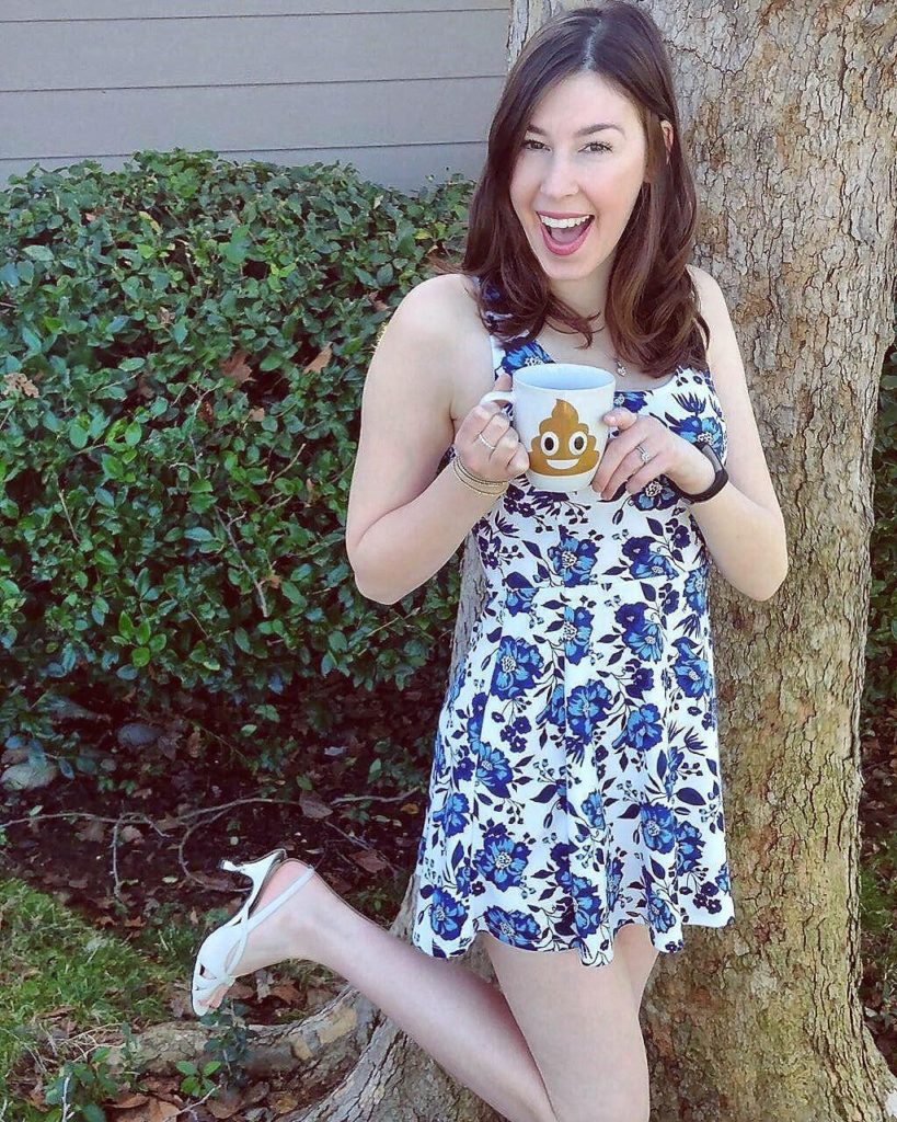 Jenna wearing blue floral dress and white hells holding poop emoji mug in front of tree